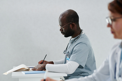 African serious doctor in uniform making notes in his notebook at table during conference