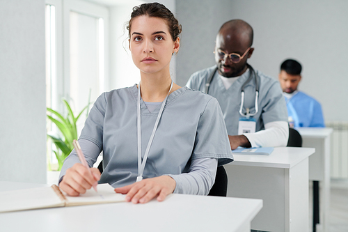 Young nurse in uniform sitting at table and making notes in notebook while listening to speaker at lecture