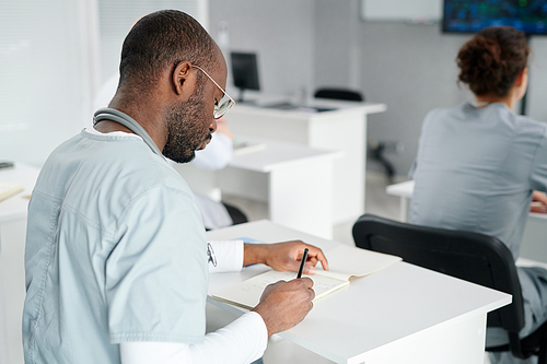 Rear view of African male doctor in uniform writing in notebook at desk during studying at class