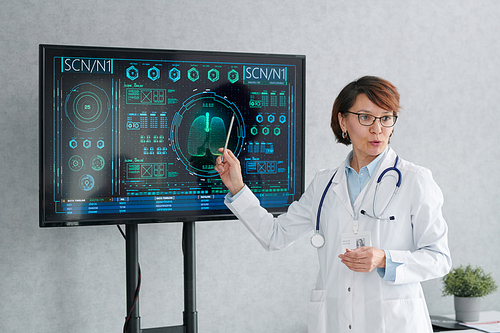 Mature female doctor in white coat pointing at digital display with chart and talking about CPR at medical training