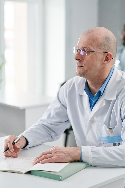 Mature doctor in eyeglasses and white coat sitting at desk with notebook during training, he focused on studying