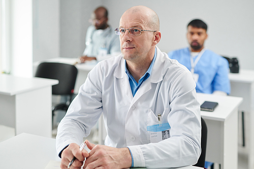 Mature male doctor in eyeglasses and white coat sitting at desk listening to lecture with serious expression