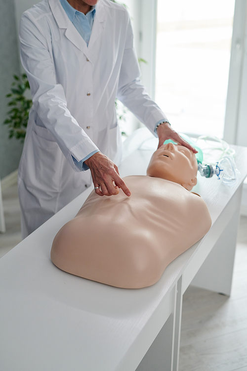 Close-up of physician in white coat pointing at chest of mannequin on table, she showing massage procedure during training