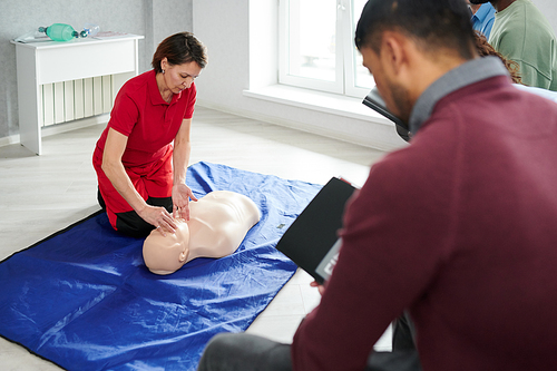 Female instructor in uniform sitting on floor with mannequin and demonstrating CPR to students during medical courses