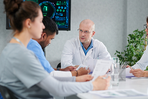 Mature male doctor in white coat sitting at table with his colleagues and discussing work during meeting at office