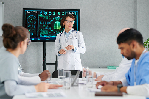 Mature female woman standing near the screen with charts and talking to her colleagues during teamwork at meeting