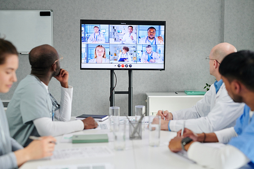 Group of doctors sitting at table and looking at big screen, they having online meeting with colleagues at office