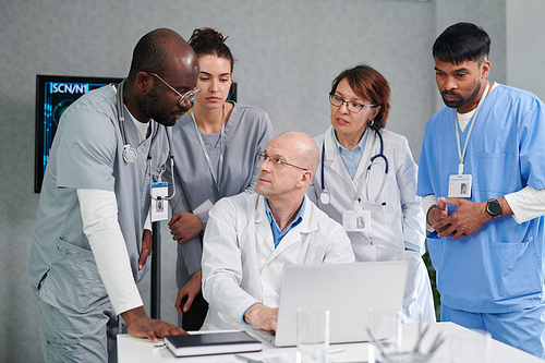 Mature male doctor sitting at table with laptop and discussing online presentation with his colleagues during teamwork at office