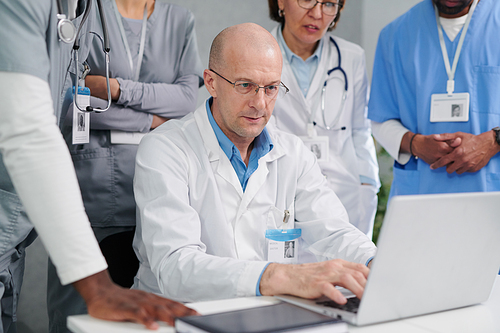 Mature male doctor in white coat showing presentation on laptop to colleagues during meeting