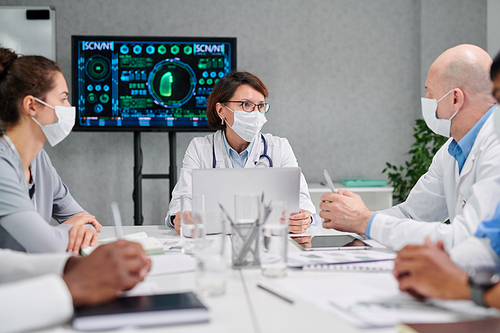 Group of doctors in mask sitting at table and discussing difficult disease together at consultation