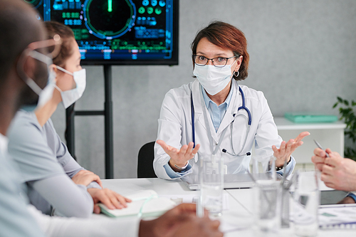 Mature female doctor in mask discussing coronavirus situation at table with colleagues during meeting