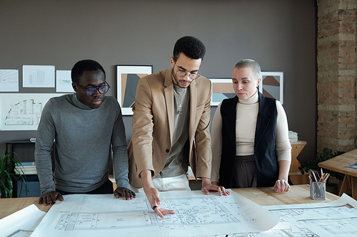 Young biracial businessman making presentation of sketch on unrolled blueprint to his colleagues at meeting