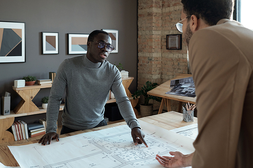 Young contemporary African architect explaining details of sketch to male colleague while pointing at blueprint