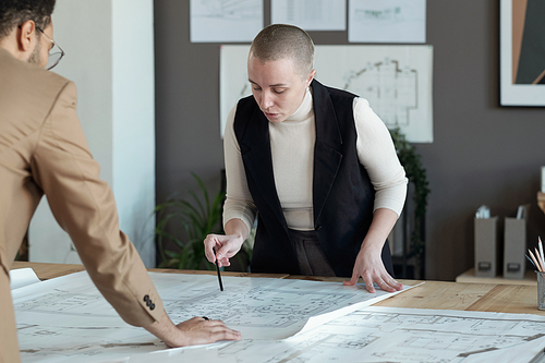 Confident businesswoman bending over table with blueprint while discussing sketch with young male colleague at meeting