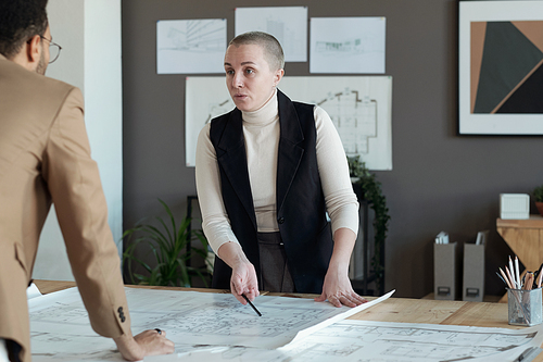 Confident mature female architect talking to male colleague while pointing at sketch on blueprint at working meeting