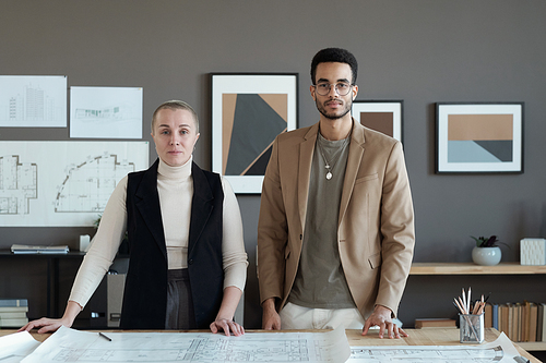 Two contemporary engineers standing by table with sketch and looking at you against grey wall with abstract paintings
