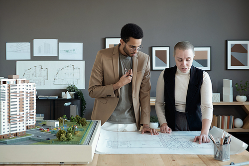 Two elegant architects or engineers discussing sketch on blueprint while standing by large table with house layout