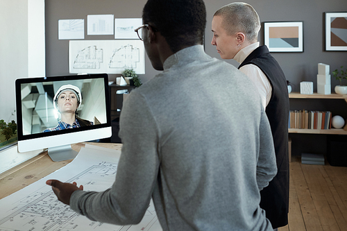 Two contemporary architects communicating to female builder on computer screen while standing by table with blueprint