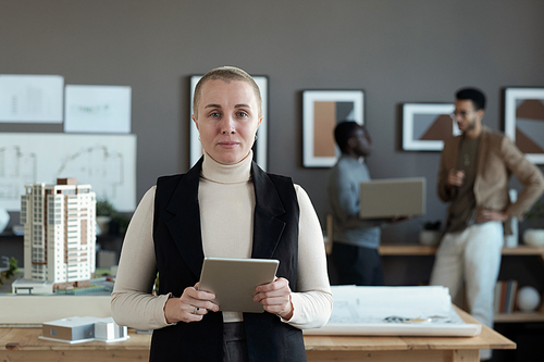 Contemporary female engineer or architect with digital tablet standing by workplace against two intercultural male colleagues