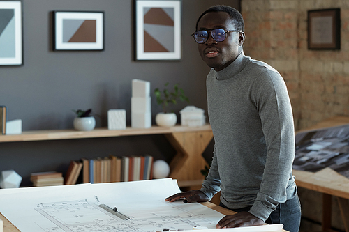 Contemporary young architect in eyeglasses and grey pullover looking at camera while standing by table with blueprint