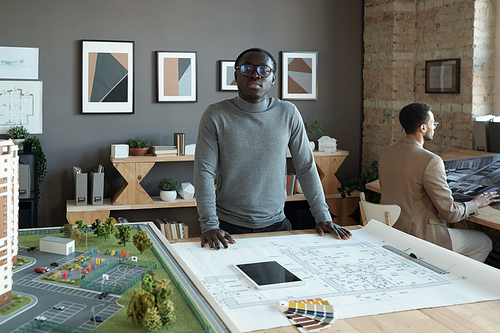 Young African engineer standing by table with sketch and house layout and looking at you while working over new project