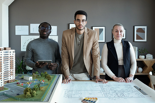 Three contemporary professionals of various ethnicities standing by table with blueprints and house layout and looking at camera