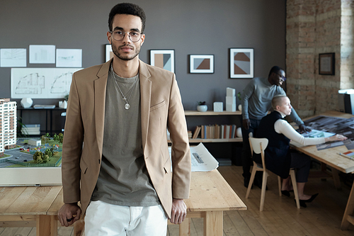 Young biracial businessman in formalwear and eyeglasses standing against wooden table with house layout and sketch