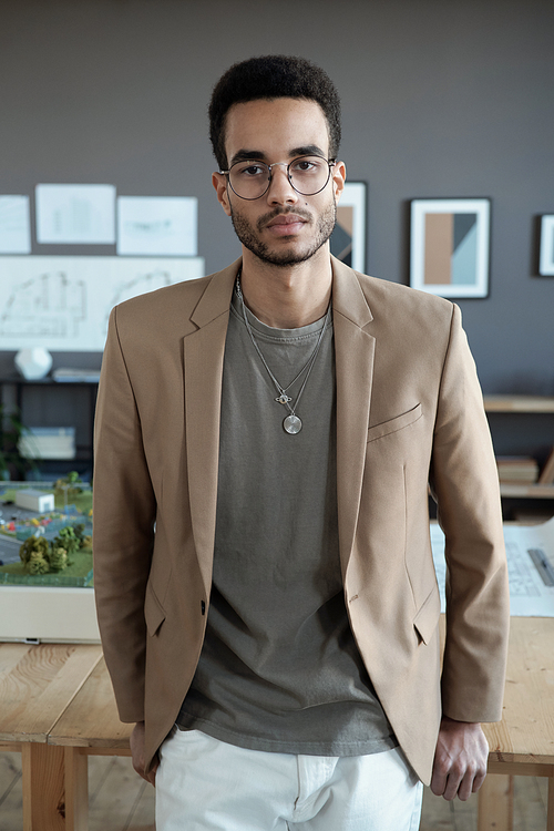 Young contemporary businessman looking at you while standing by his workplace against office environment