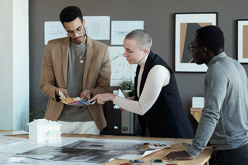 Three colleagues choosing colors for apartment interior from palette held by young biracial male designer at working meeting