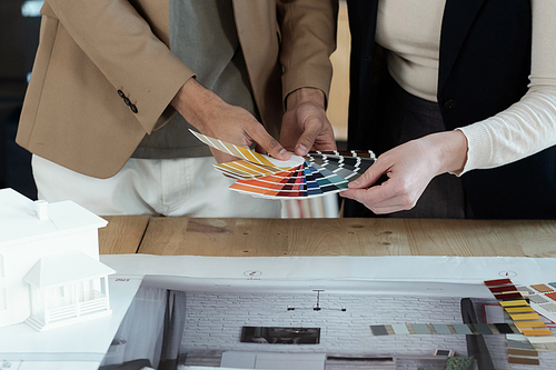 Hands of two designers of interior choosing color for apartment walls while standing by table with blueprint and photos