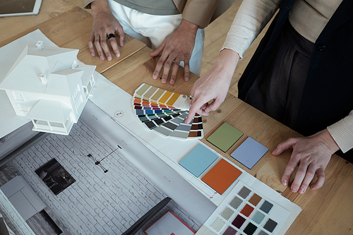 Hand of businesswoman pointing at color palette while consulting with male colleague over table with blueprint, photos and house layout