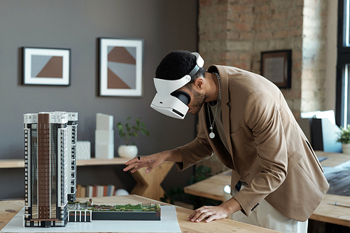 Young architect in vr headset bending over house layout while standing by table and making presentation of new architectural project