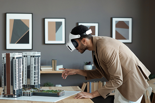 Young biracial businessman in vr headset bending over table and pointing at house layout while making virtual presentation