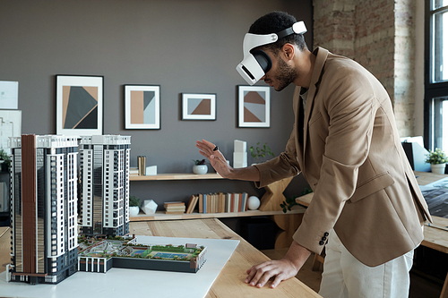 Young elegant architect in vr headset standing by table with house layout during presentation of his new project