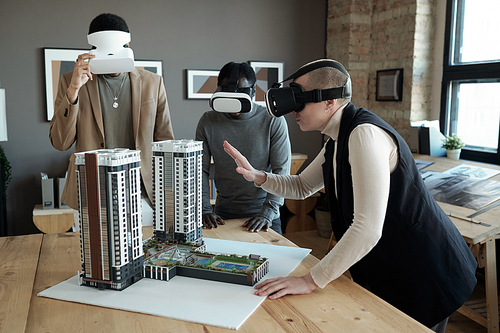 Group of three architects in vr headsets standing by table with layout of new block of flats while one of them making presentation