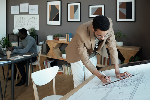 Young biracial architect in smart casualawear drawing line on sketch while bending over workplace against African male coworker
