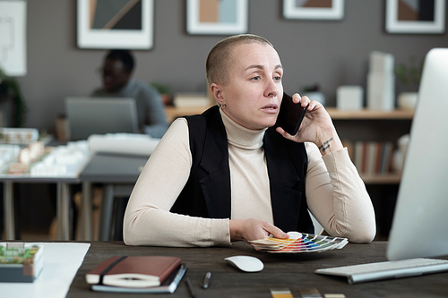 Contemporary female designer speaking on mobile phone while sitting in front of computer monitor and consulting clients