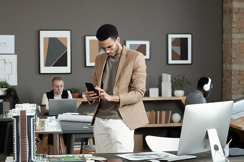 Young biracial businessman scrolling in smartphone while standing by workplace with computer monitor and house layout