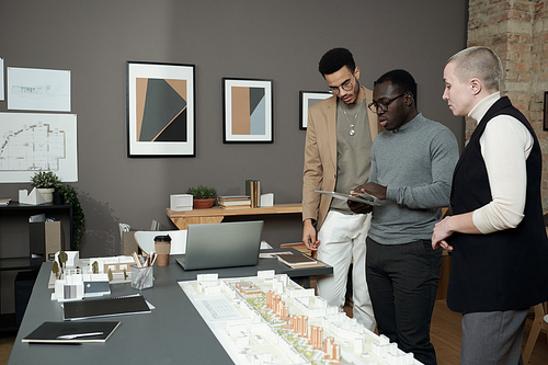 Three creative designers or architects discussing layout at meeting while African man with tablet making presentation
