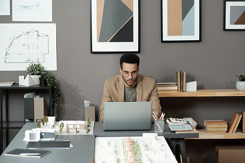 Young serious biracial businessman looking at laptop screen while sitting by table with layouts and other office supplies
