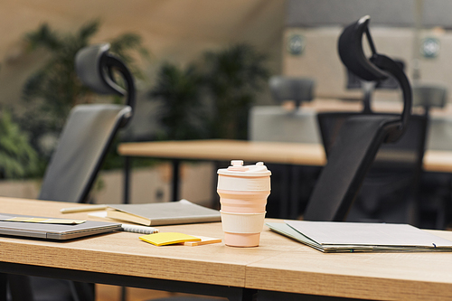 Close up background image of modern open space office decorated with plants, focus on coffee cup on wooden table in foreground, copy space