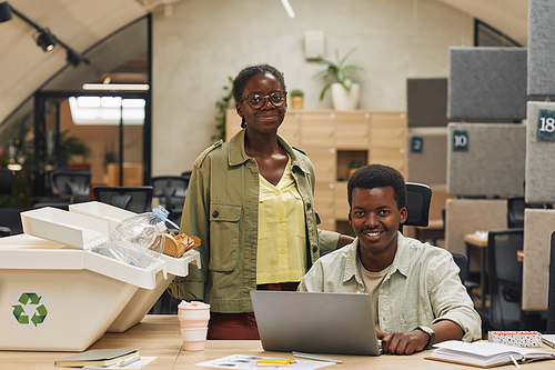Portrait of smiling African-American people looking at camera while posing by waste sorting bins in modern office, copy space