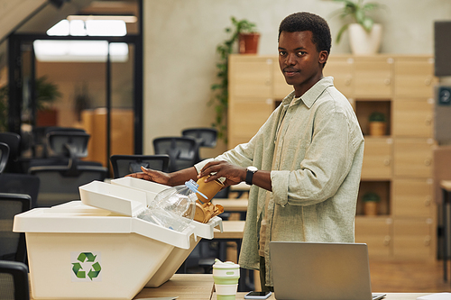 Side view portrait of young African-American man putting paper cup into waste sorting bin in office, copy space