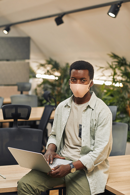 Vertical portrait of young African-American man wearing mask and looking at camera while using laptop in post pandemic office
