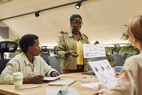 Portrait of young African-American woman holding data chart while giving report of statistics and performance during business meeting in modern office, copy space