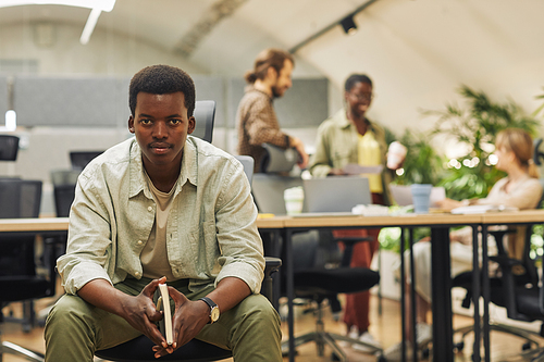 Portrait of young serious African-American man looking at camera while sitting in modern office with people working in background, copy space