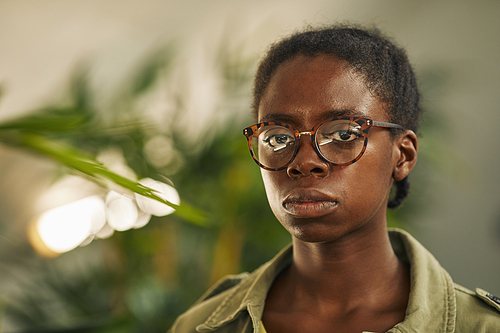 Head and shoulders portrait of contemporary African-American woman wearing glasses and looking at camera while standing in modern office with plants, copy space