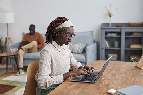 Side view portrait of young African-American woman using laptop at desk while working from home in cozy apartment interior, copy space