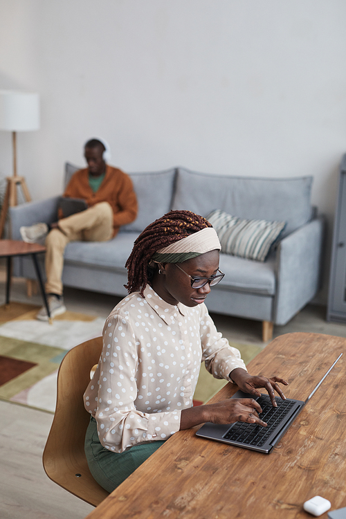 Vertical high angle portrait of young African-American woman using laptop at desk while working from home in cozy apartment interior