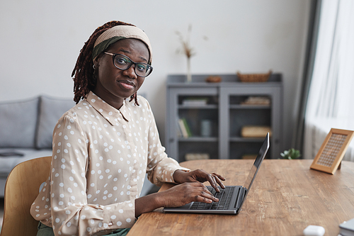 Portrait of young African-American woman using laptop at desk and looking at camera while working from home in apartment interior, copy space